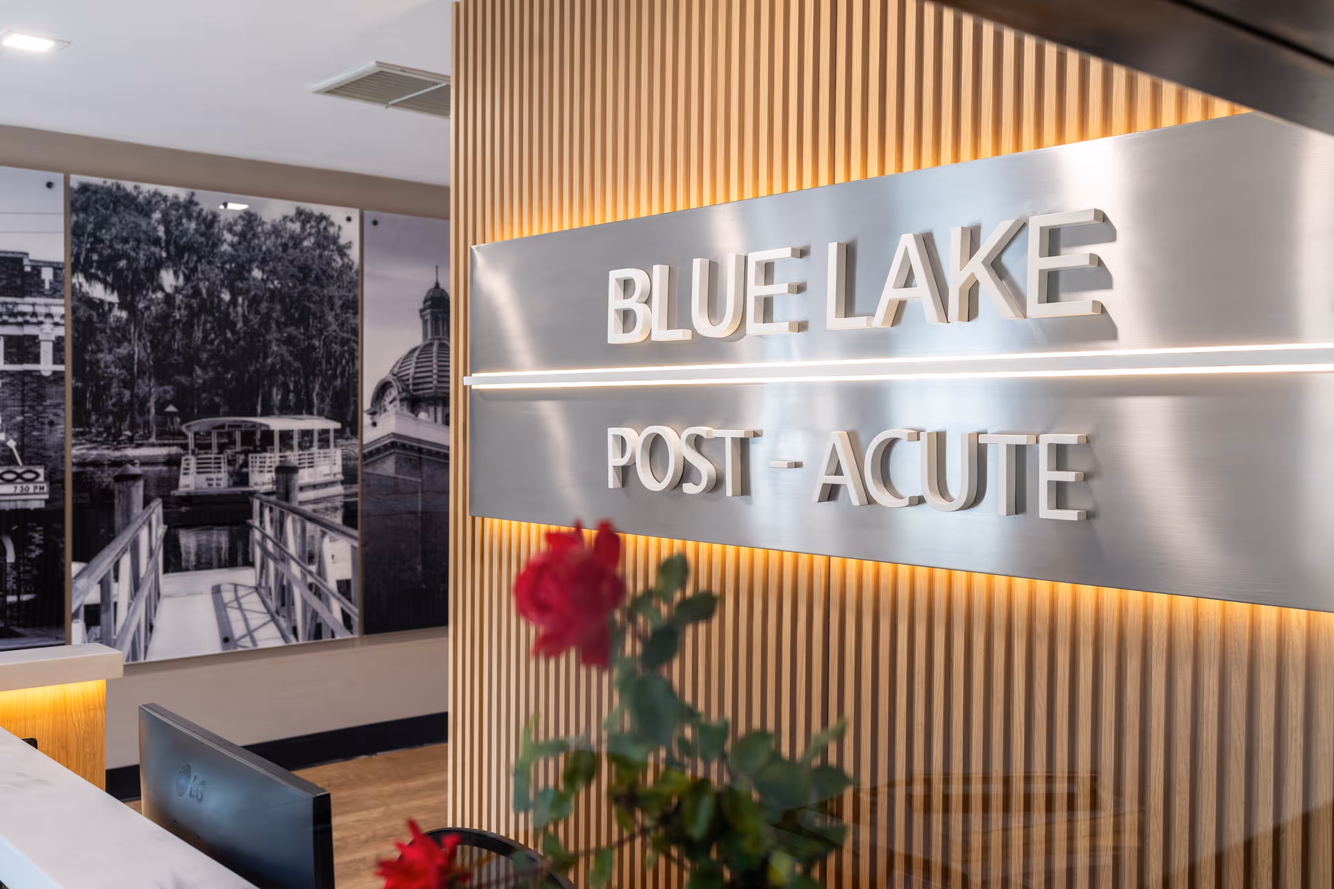 Reception area of Blue Lake Post Acute facility with a metallic sign displaying the facility name on a wooden slat wall. A red flower plant is visible in the foreground, and a black and white photo mural of a dock and trees is on the adjacent wall.
