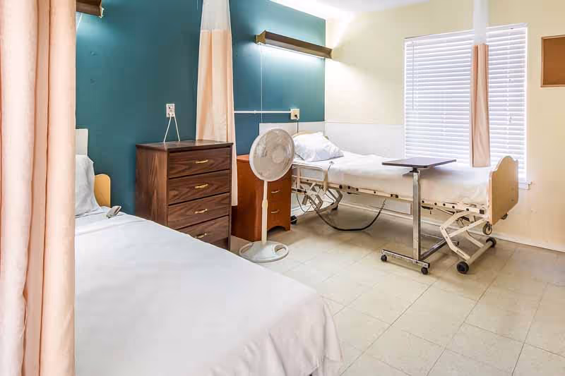 A nursing facility room with two hospital beds separated by curtains. Each bed has white bedding and pillows. There is a wooden dresser between the beds, a standing fan, and a small rolling table near one bed. The walls are painted teal and beige, and a window with white blinds lets in natural light.