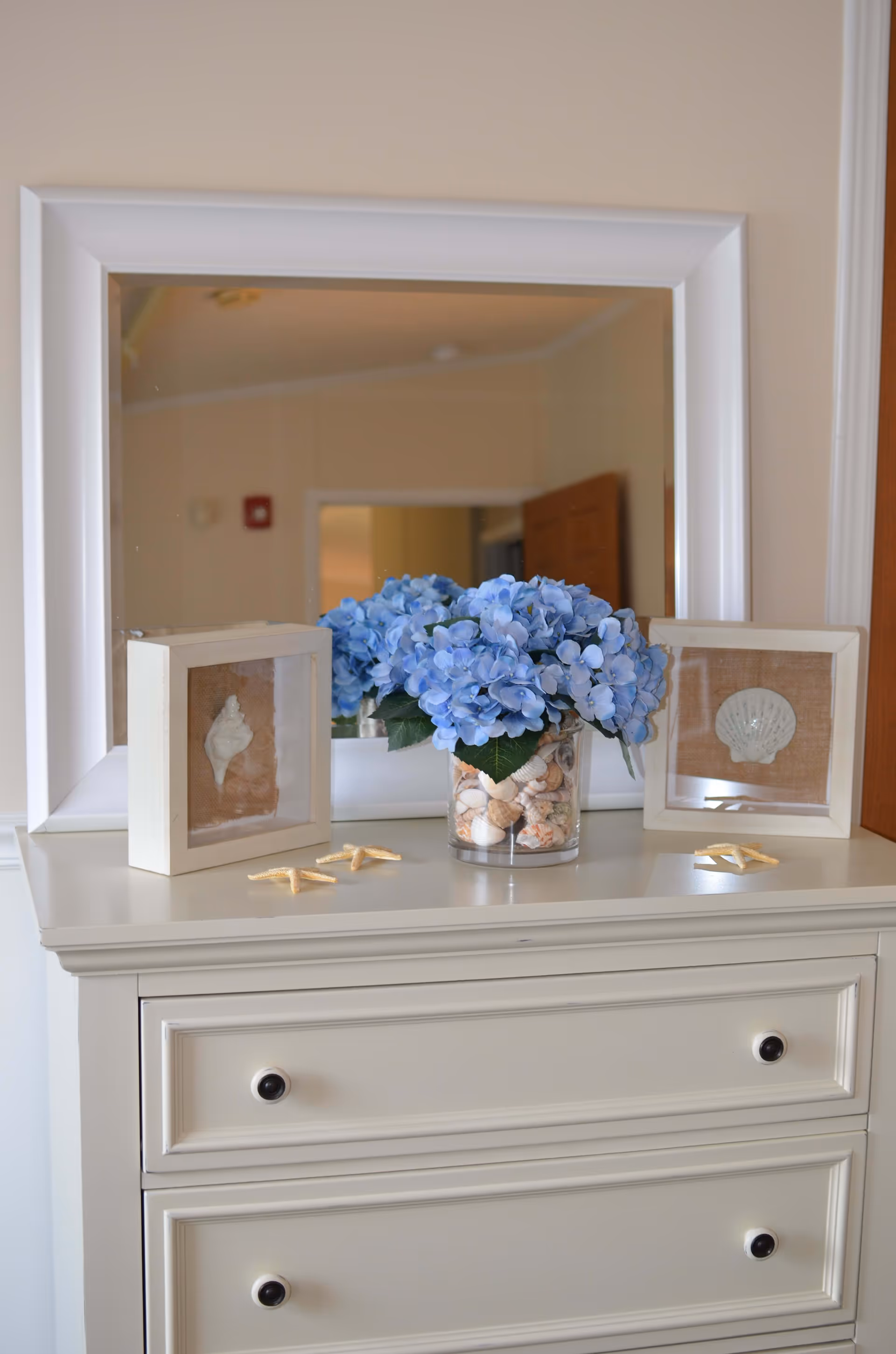 White dresser topped with a mirror, a vase of blue hydrangeas and decorative shell-framed accents.