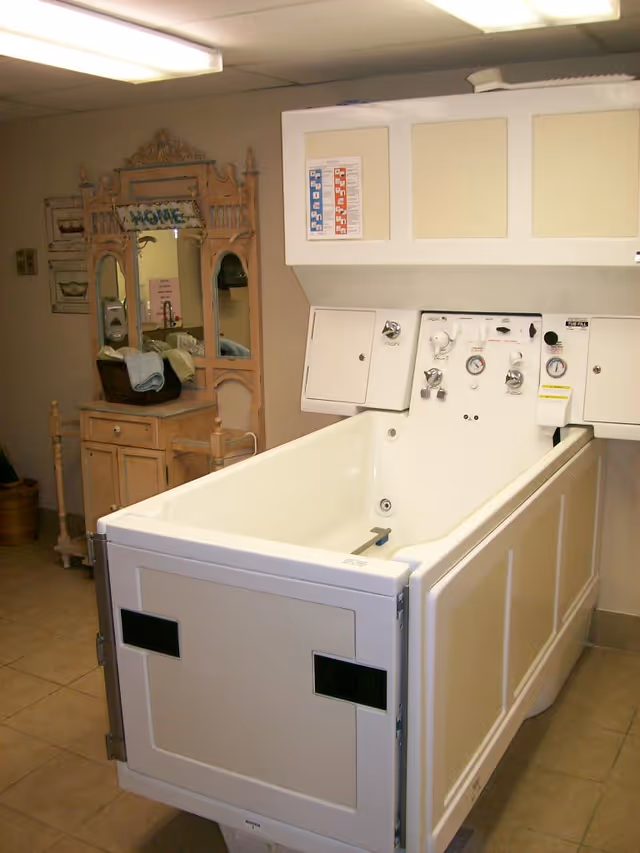A specialized bathing tub with control panels and gauges, located in a room with tiled floors and beige walls. In the background, there is a wooden vanity with a mirror and a basket containing towels. The room is softly lit by ceiling lights.