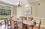 A dining room with a wooden dining table set for six people. The table has white plates, cups, and silverware. There are six chairs with patterned upholstery around the table. A large window on the left side lets in natural light and shows a green outdoor area. A decorative framed artwork hangs on the beige wall, and a chandelier with multiple lights hangs above the table.