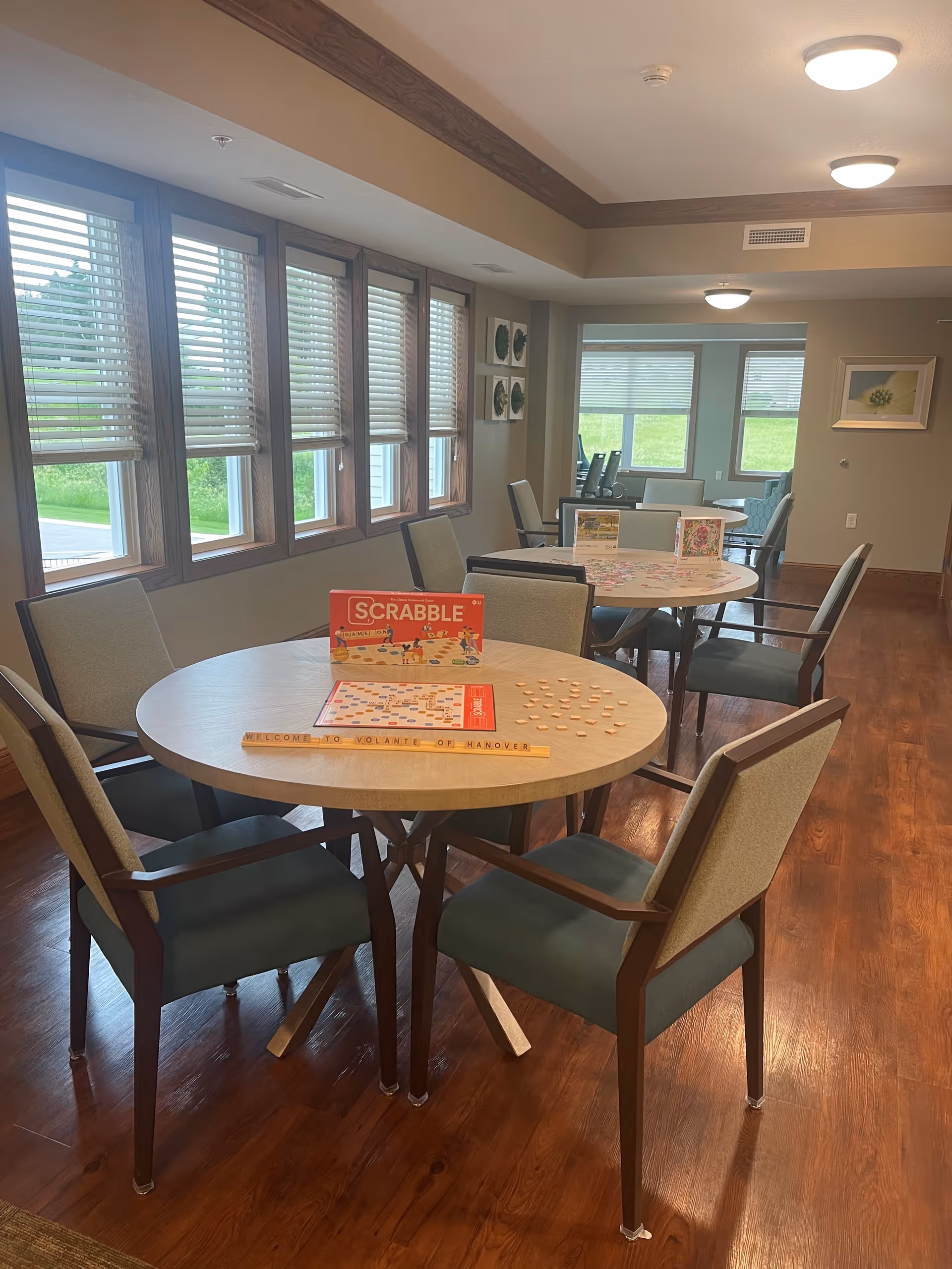 A bright room with wooden floors and multiple round tables surrounded by chairs. One table in the foreground has a Scrabble game set up with tiles and a board, along with a wooden tile holder that reads 'WELCOME TO VOLANTE OF HANOVER'. Large windows with blinds line one wall, letting in natural light. The room appears to be a common area or activity room in a senior living facility.