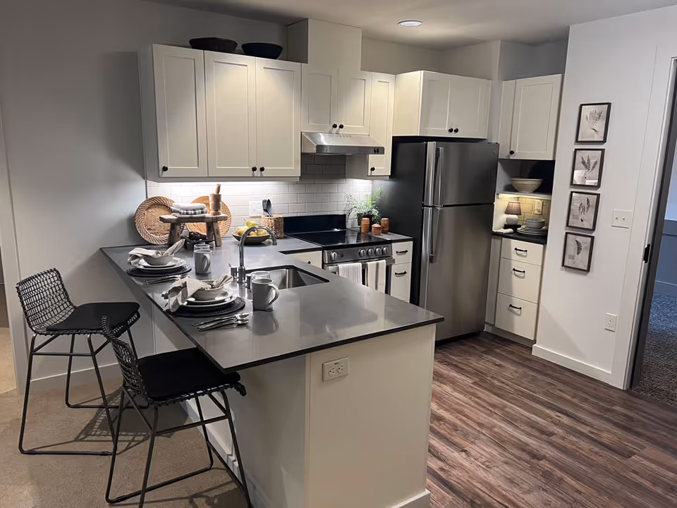 Modern furnished kitchen with an island counter and bar stools, stainless steel refrigerator and stove, and white cabinets.