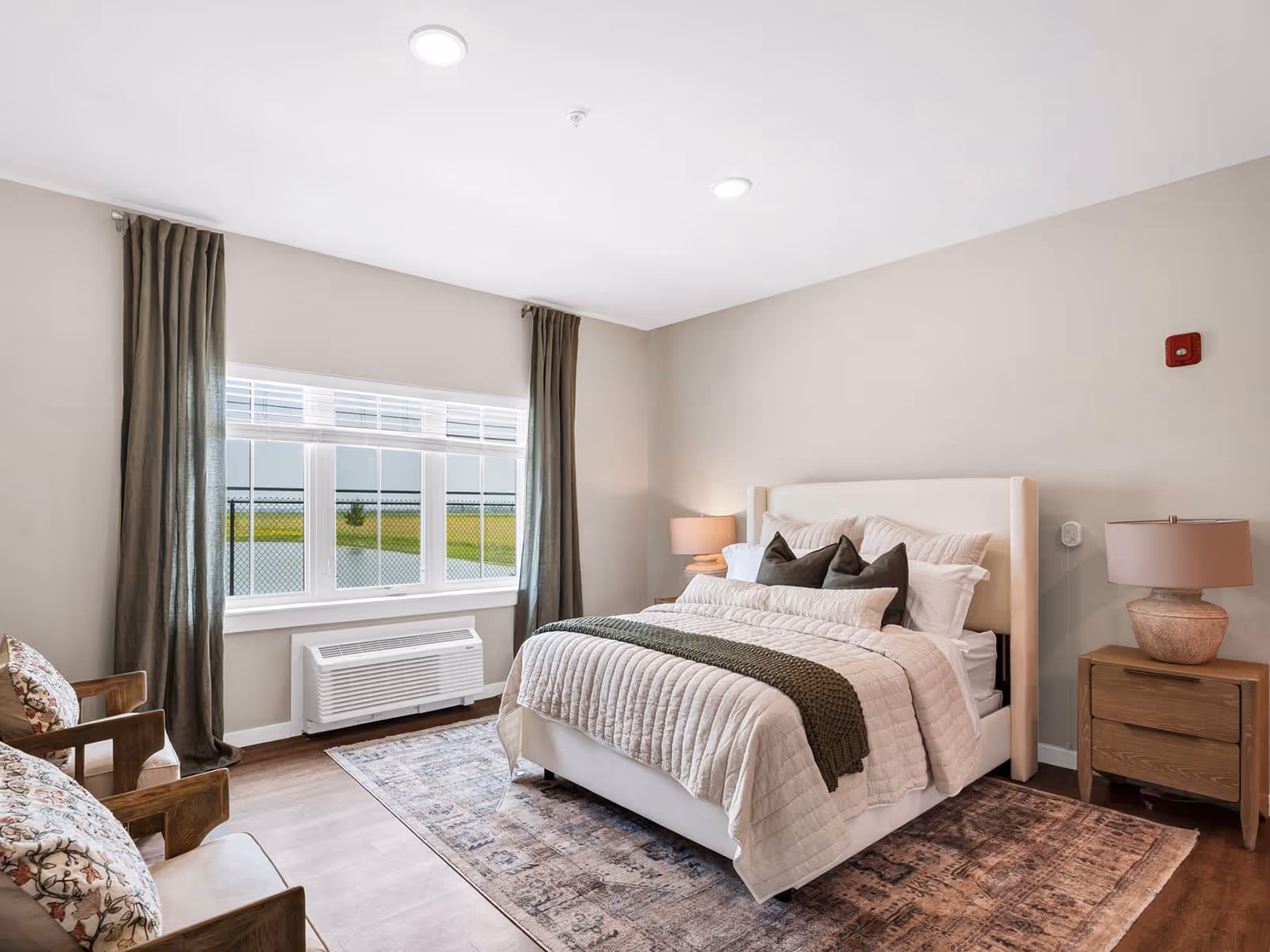 A bright and cozy bedroom in a senior living facility featuring a neatly made bed with multiple pillows and a green throw blanket. The room has a large window with dark curtains, two wooden chairs with floral cushions, a wooden nightstand with a lamp, and a patterned area rug on a wooden floor.