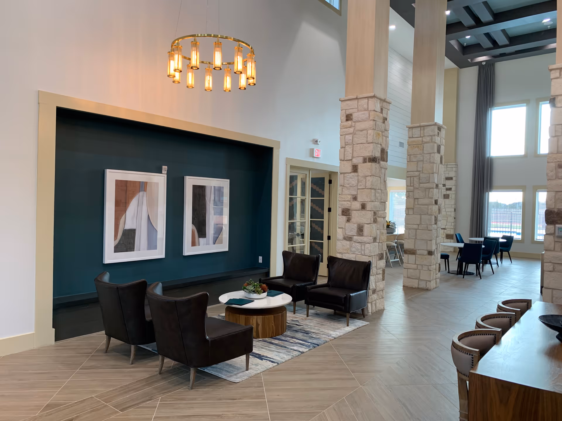 A modern and spacious common area with high ceilings and large windows allowing natural light. The room features stone pillars, a seating area with four dark leather chairs around a small round table on a rug, and a wooden table with several chairs. Two abstract framed artworks hang on a dark green accent wall, and a circular chandelier with multiple lights hangs from the ceiling.