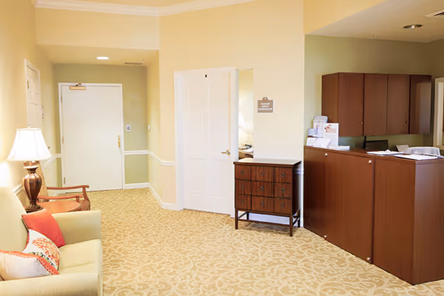 A well-lit interior space with beige walls and patterned carpet. On the left, there is a green sofa with colorful pillows and a wooden chair next to a lamp on a side table. In the center, there is a white door and a small wooden chest of drawers. On the right, there is a wooden reception desk with papers and office supplies on top.