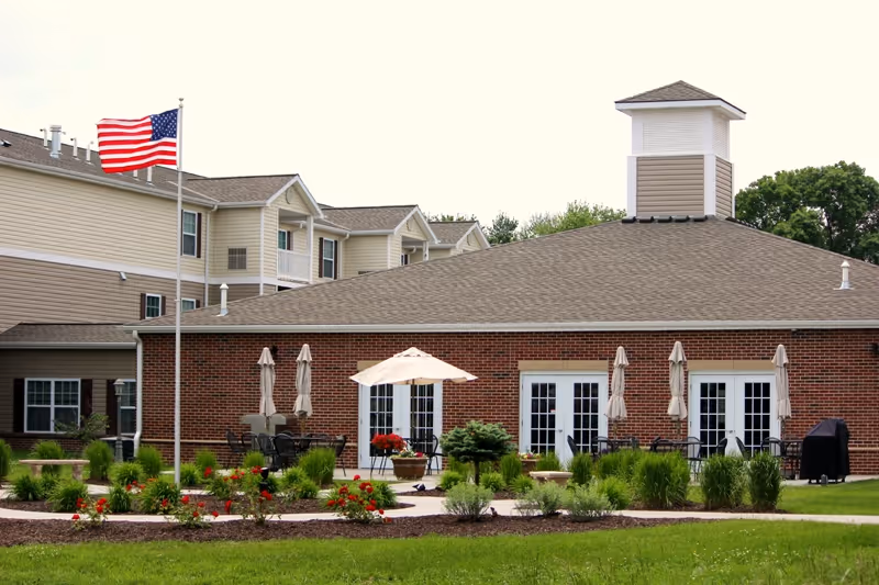 Exterior view of a senior living facility with a brick building featuring multiple glass doors, outdoor patio tables with umbrellas, an American flag on a flagpole, and well-maintained landscaping with green shrubs and red flowers.