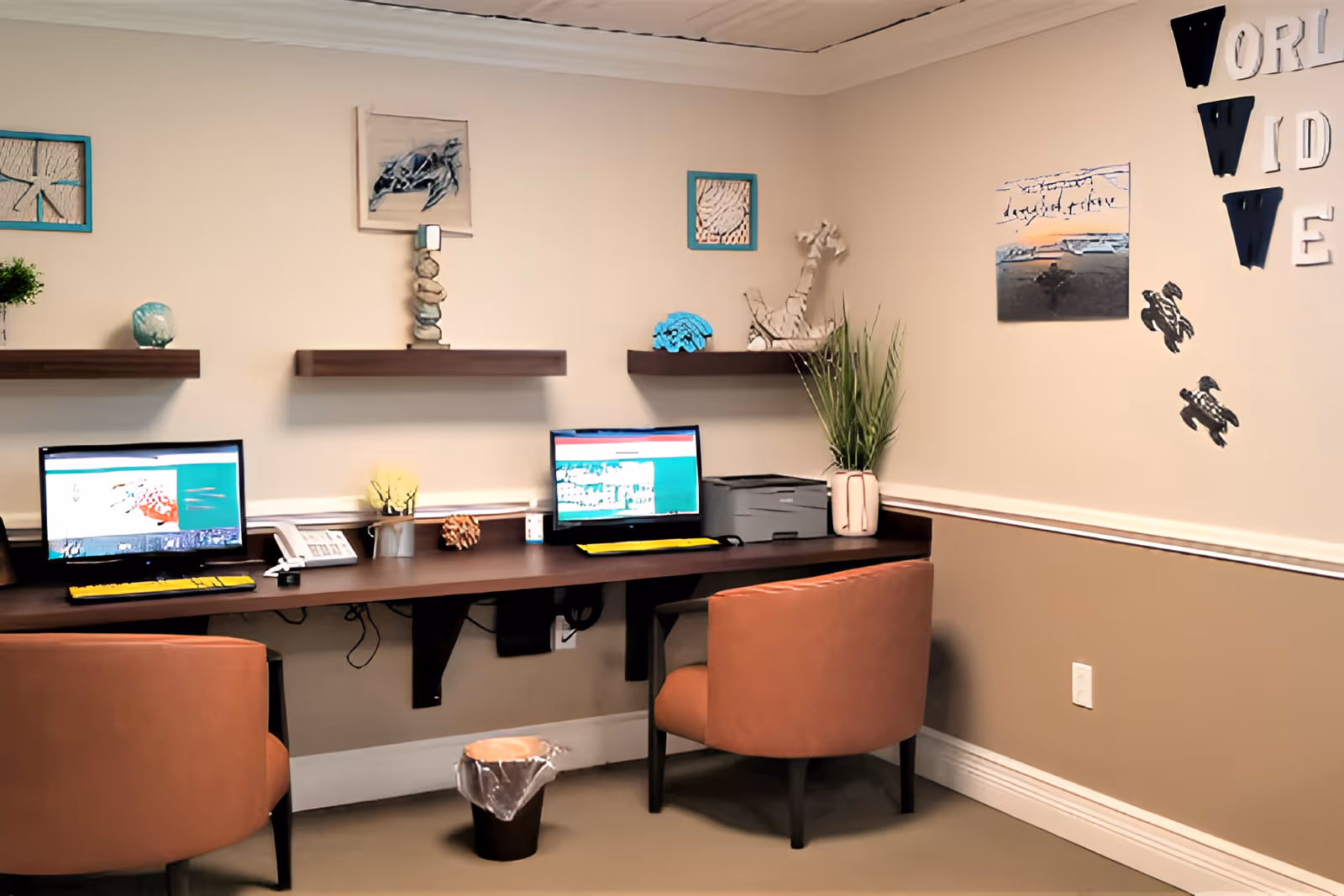 A small office or computer workstation area with two brown chairs facing a long wooden desk mounted to the wall. Two computer monitors with keyboards are on the desk, along with a telephone and a printer. The beige walls are decorated with small shelves holding decorative items and framed pictures. On the right wall, there are wall decorations including the words 'WORLD WIDE' and images of turtles.