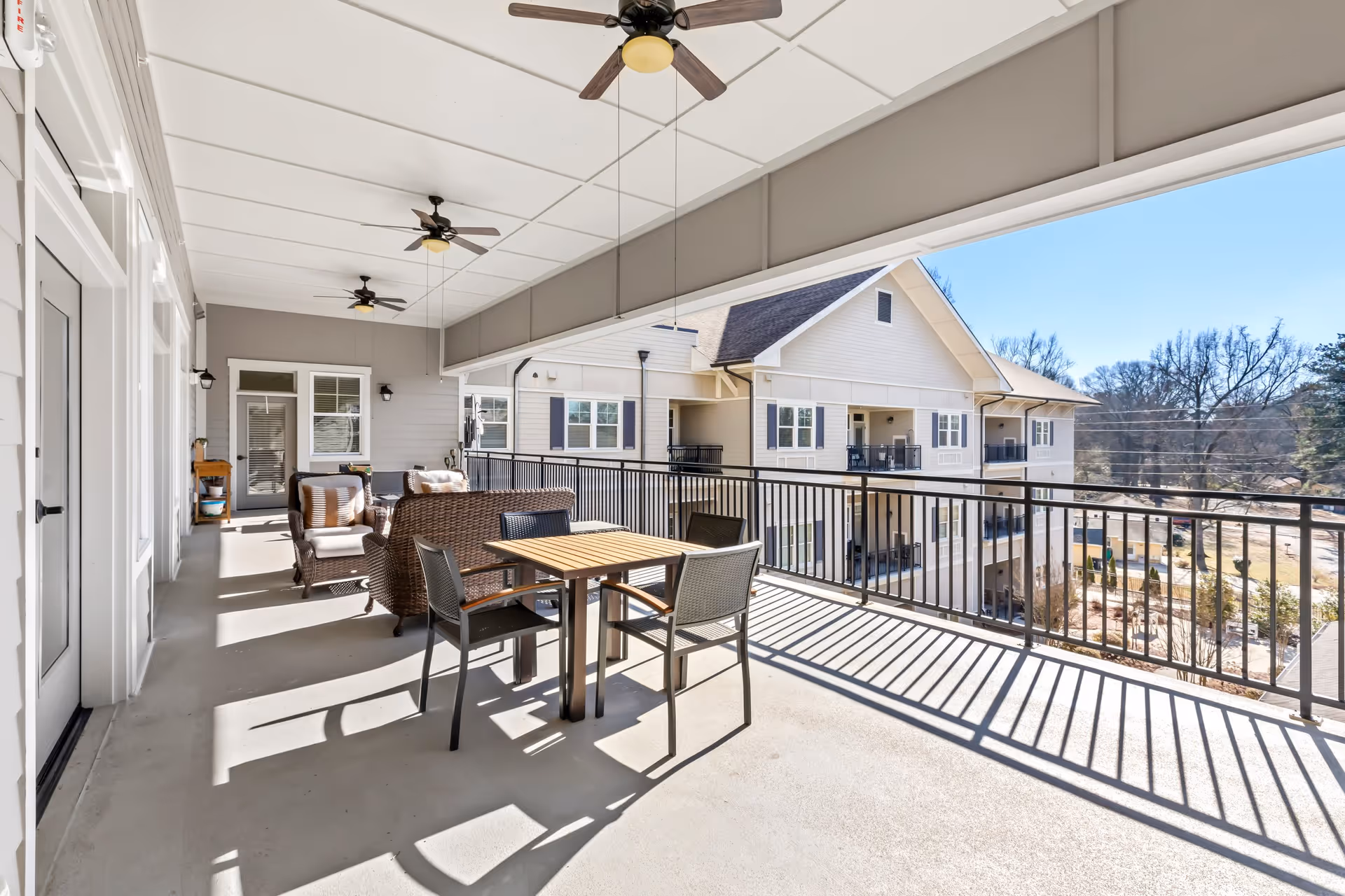 A spacious covered balcony with ceiling fans, outdoor seating including a table with four chairs and a wicker sofa set. The balcony overlooks a residential building and a landscaped area with trees under a clear blue sky.