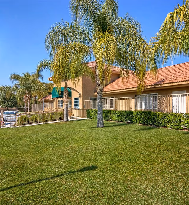 Exterior view of Desert Hills Memory Care Center showing a beige building with a red tile roof, green awnings, palm trees, and a well-maintained grassy lawn under a clear blue sky.