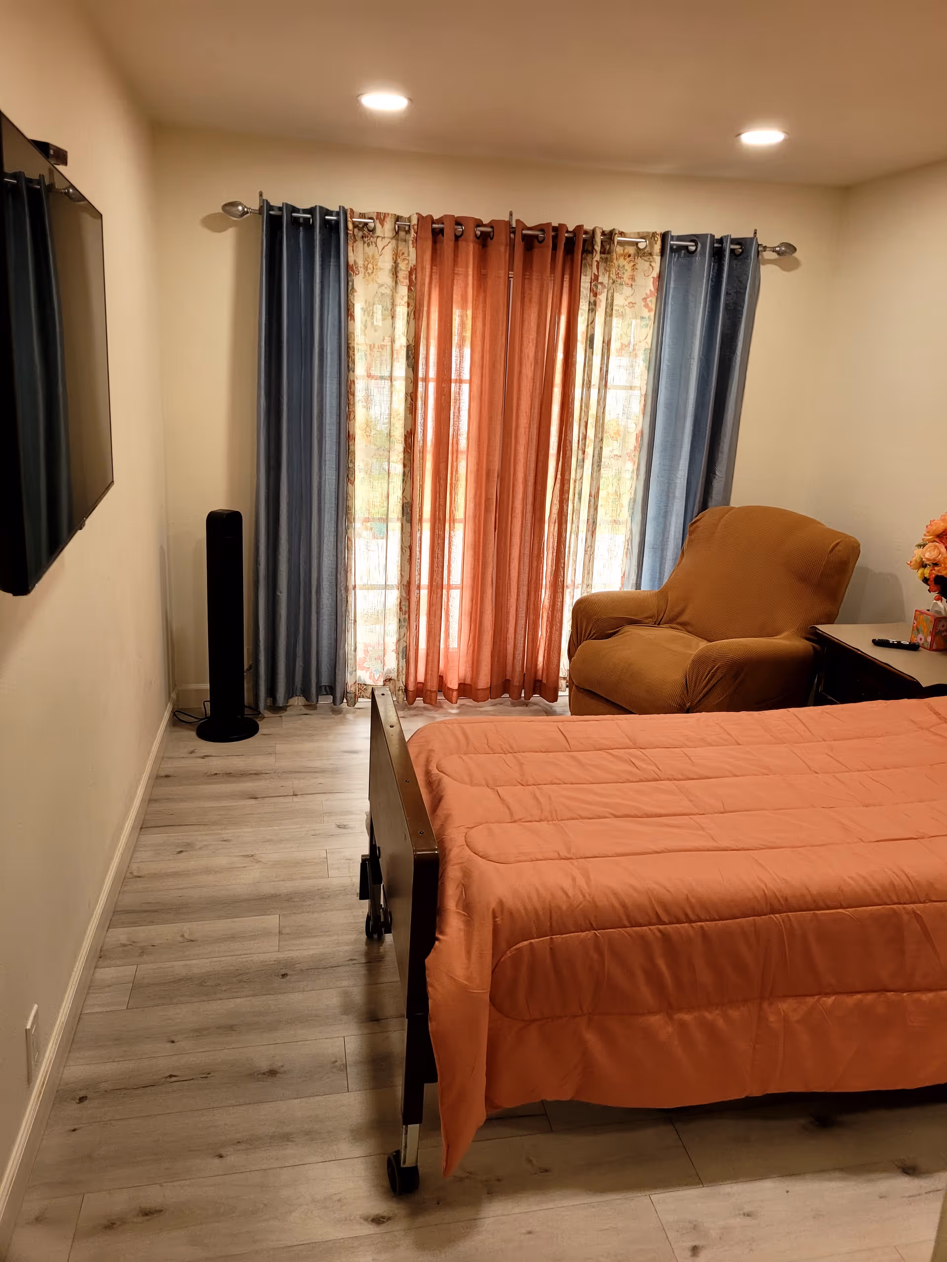 A bedroom with a single bed covered in an orange quilt, a brown armchair, a side table with a flower arrangement, and a wall-mounted flat screen TV. The room has light wood flooring and a window with blue, floral, and orange curtains.