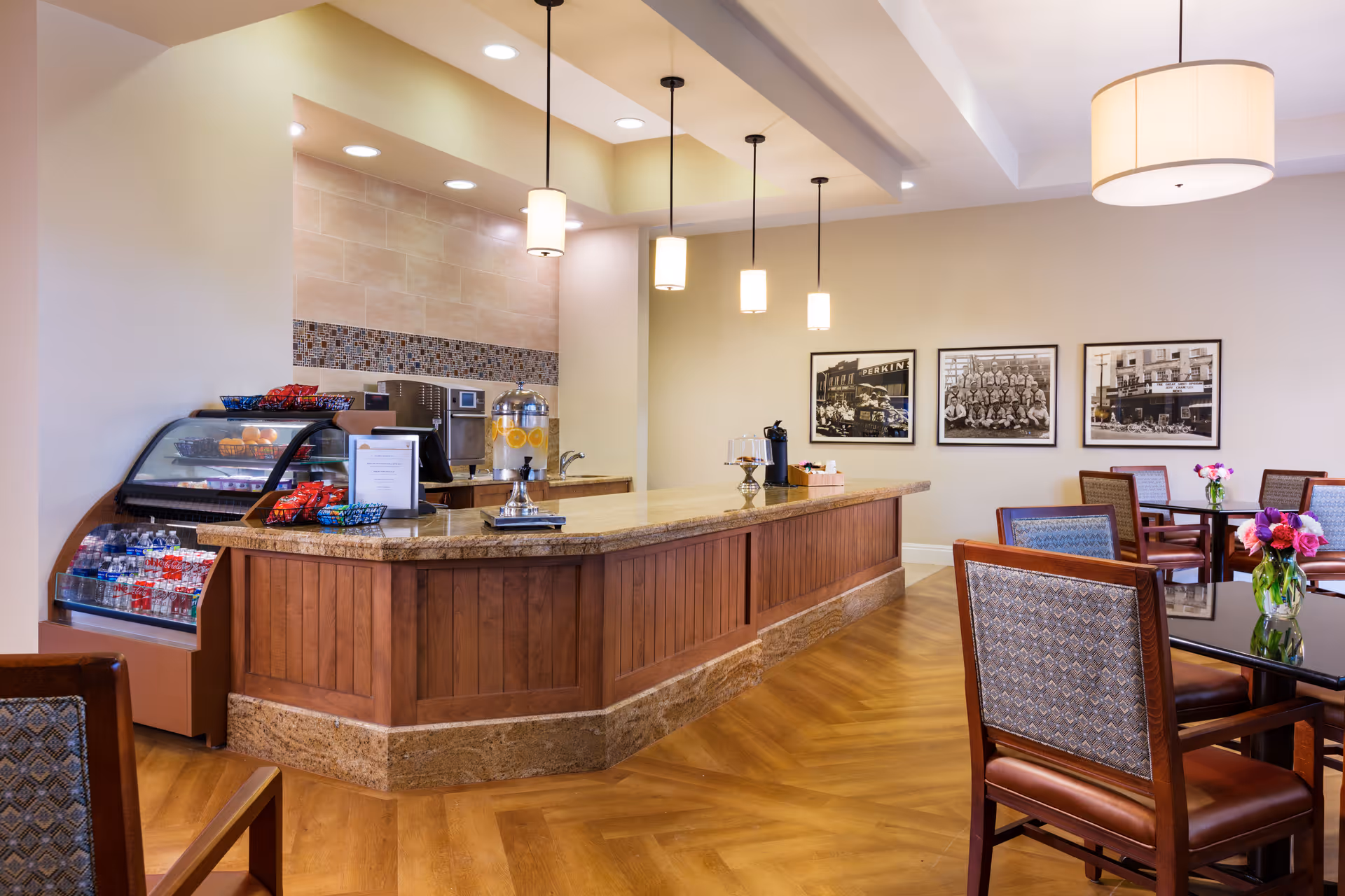 Interior view of a dining area with a wooden counter displaying snacks, a beverage dispenser, and a coffee machine. There are several tables and chairs with patterned upholstery, fresh flowers on the tables, and framed black-and-white photographs on the wall. The space is well-lit with pendant lights and recessed ceiling lights.