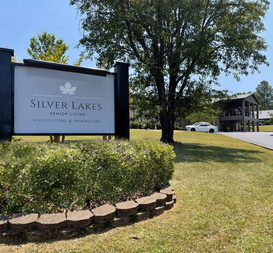 A Silver Lakes Senior Living entrance sign on a grassy lawn with trees and the facility building and a parked car in the background.