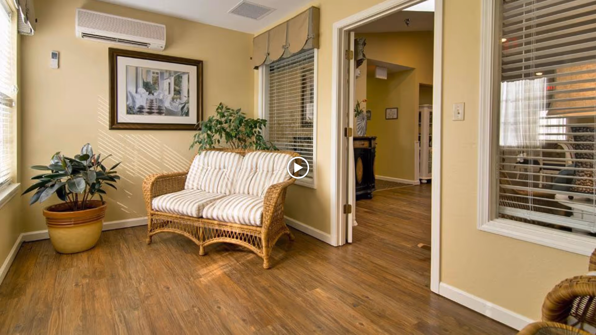 A cozy sitting area in a senior living facility with a wicker loveseat featuring striped cushions, two potted plants, a framed painting on the wall, and windows with blinds letting in natural light. The room has wooden flooring and beige walls, with an open doorway leading to another room.