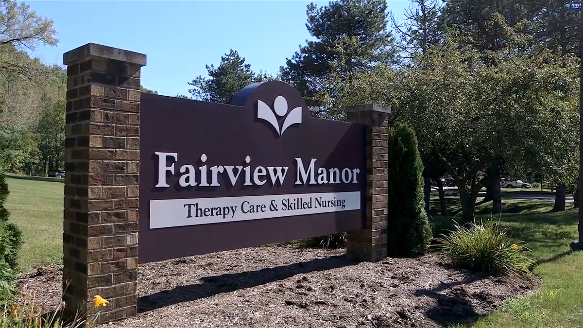 Outdoor view of a large sign for Fairview Manor, a therapy care and skilled nursing facility, set between two brick pillars with trees and grass in the background.