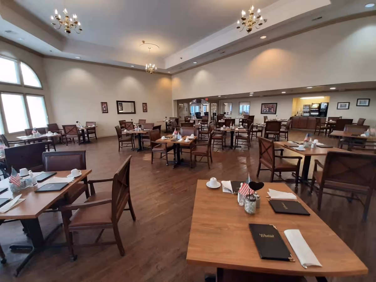 Spacious dining room with multiple wooden tables and chairs arranged neatly. Each table is set with menus, napkins, cups, and small American flags. The room has large windows allowing natural light, chandeliers hanging from the ceiling, and a beverage station in the back corner.