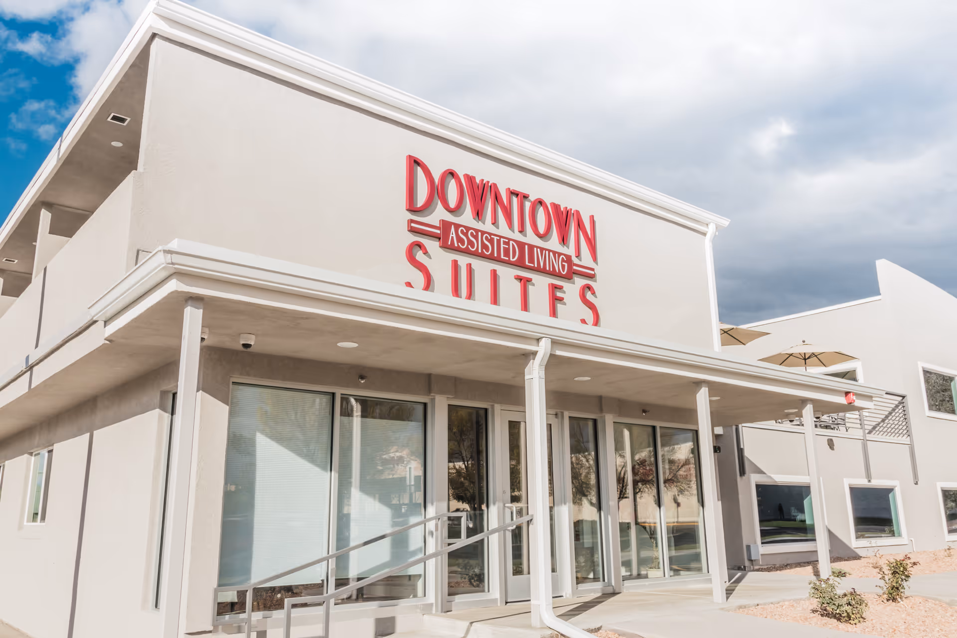 Exterior view of a modern assisted living facility building with large glass doors and windows, a covered entrance with a ramp, and a sign that reads 'Downtown Assisted Living Suites' in red letters.