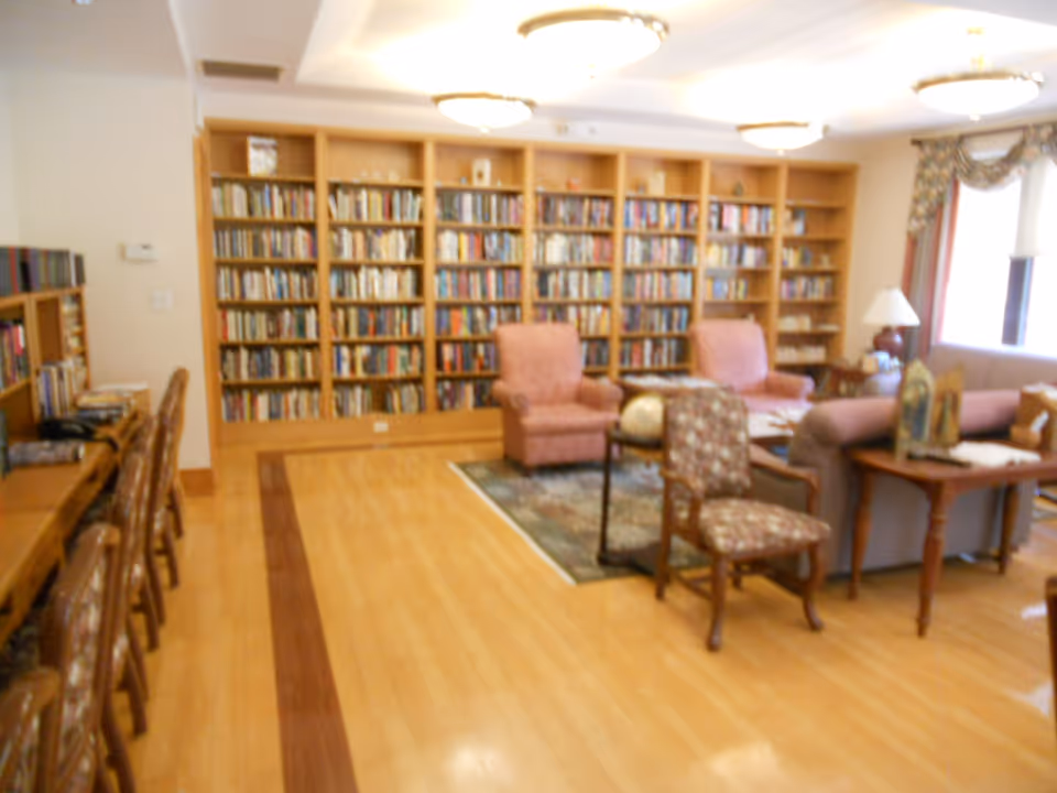 A cozy library or reading room with wooden bookshelves filled with books along the back wall. There are several upholstered chairs and a sofa arranged around a small table on a rug. To the left, there is a long wooden desk with chairs. The room has wooden flooring and is well-lit with ceiling lights and natural light from windows with curtains.