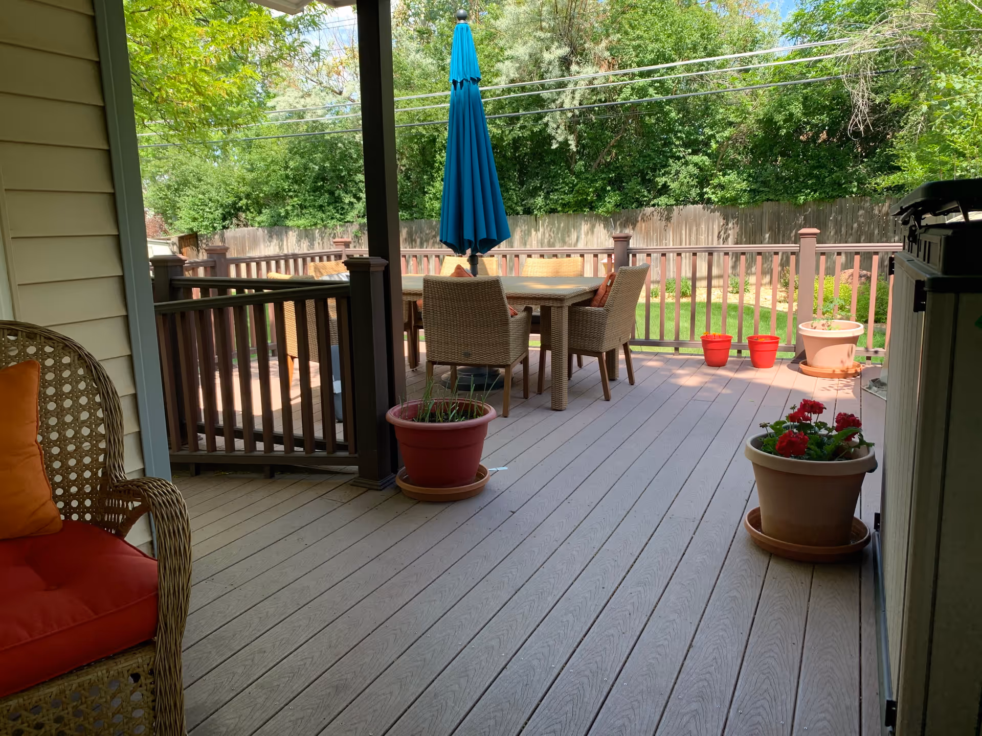 A wooden outdoor deck with a table and four wicker chairs under a closed blue patio umbrella. There are several potted plants on the deck, including red flowers and green plants. A wicker chair with red cushions is partially visible on the left side. The deck is surrounded by a wooden railing and overlooks a green backyard with trees and a wooden fence.