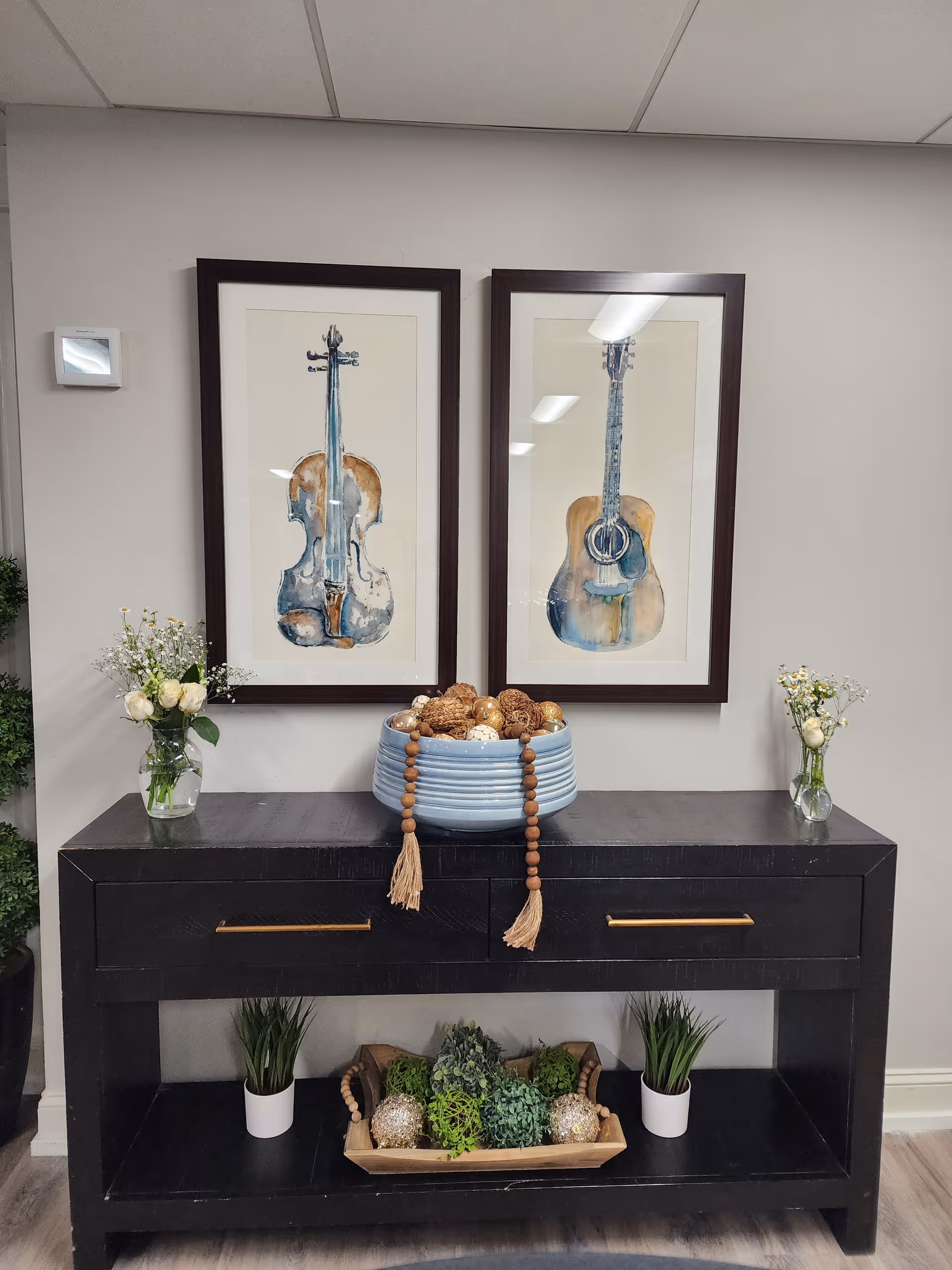 A black wooden console table with two drawers and an open shelf at the bottom. On top of the table, there is a large blue bowl filled with decorative balls and a wooden bead garland draped over the bowl. On each side of the bowl, there are small glass vases with white flowers and greenery. Below on the open shelf, there are two small potted plants and a wooden tray with green and gold decorative balls. Above the table, two framed watercolor paintings of musical instruments, a violin and an acoustic guitar, hang on a light gray wall.