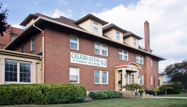 Exterior view of a large brick building with multiple windows and a small porch entrance. A sign on the building reads 'CELEBRATING A Century of Caring.' The building is surrounded by green bushes and a lawn under a partly cloudy sky.