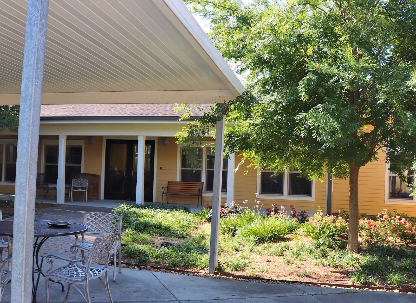 Outdoor courtyard with metal patio tables and chairs, a covered walkway, landscaped garden, and the yellow exterior of the facility.