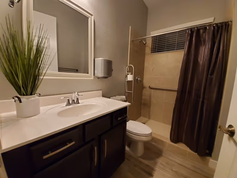 A clean bathroom featuring a white sink with a silver faucet set in a dark wood vanity with drawers and cabinets. Above the sink is a large rectangular mirror with a white frame. To the right, there is a toilet and a walk-in shower with beige tiles, a silver grab bar, and a dark brown shower curtain. A small window with blinds is above the shower. A white pot with tall green grass-like plants is placed on the vanity countertop.