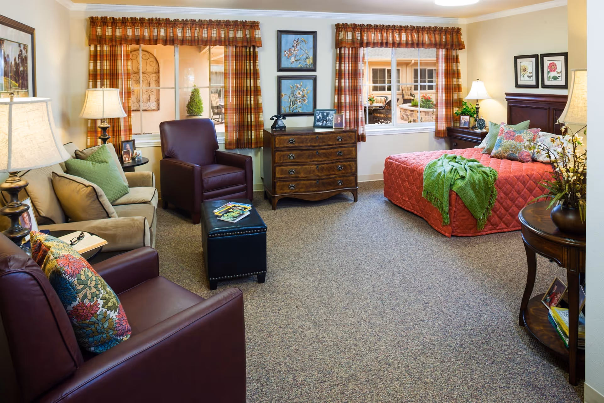A cozy bedroom in a senior living facility featuring a bed with a red quilt and green throw, two windows with plaid curtains, a wooden dresser with framed pictures and decorative items, two leather armchairs with colorful pillows, a beige sofa with green pillows, a small round table with a lamp, and a round side table with flowers and books.