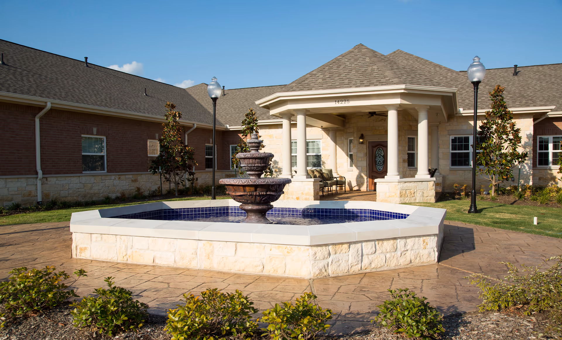 Exterior view of a senior living facility named The Heritage Communities featuring a stone and brick building with a covered entrance supported by columns. In front of the entrance is a large octagonal stone fountain with water, surrounded by a paved area and landscaping with bushes and small trees. Two street lamps are visible near the entrance.