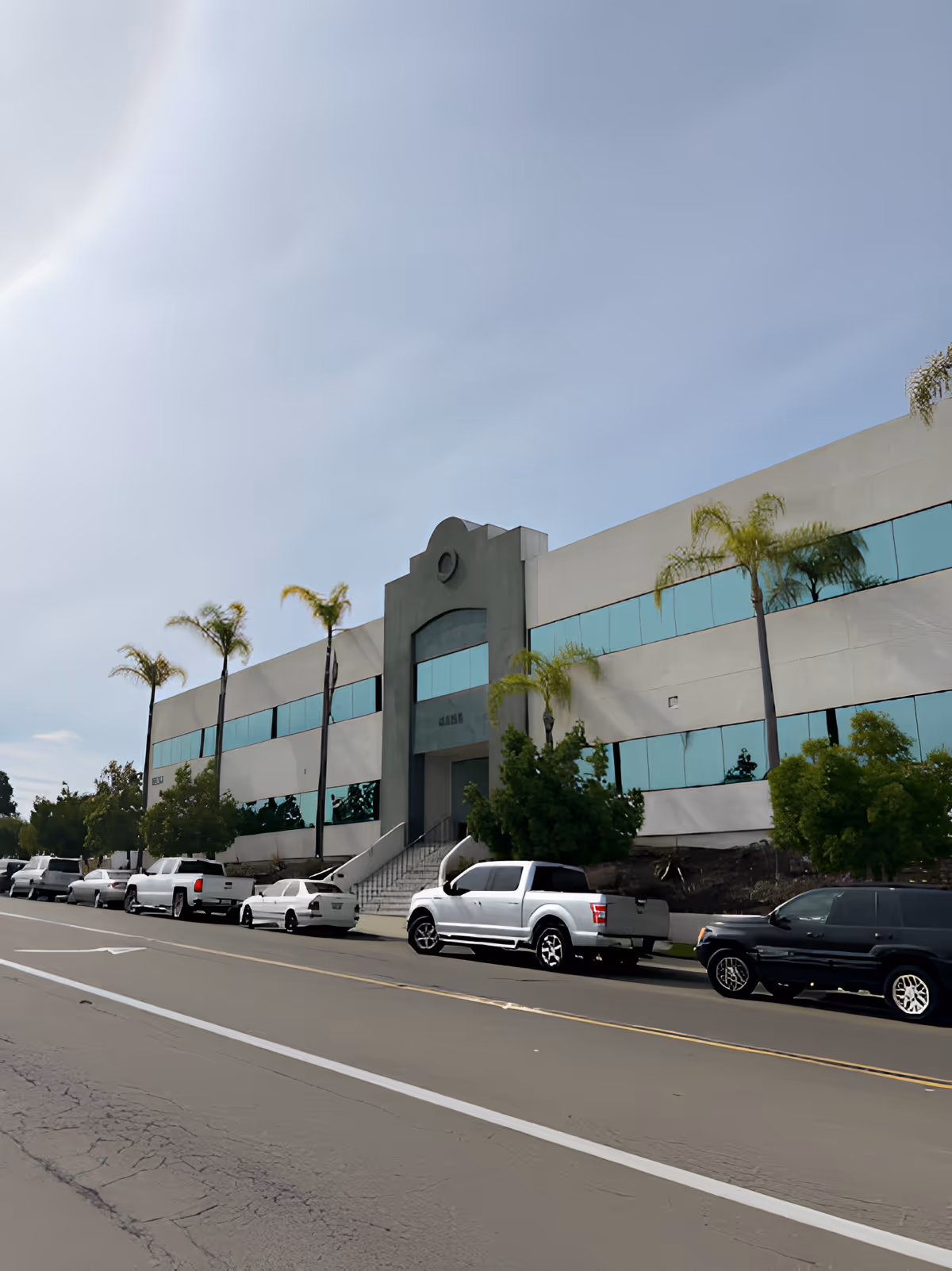 Exterior view of a two-story commercial building with large reflective windows and palm trees lining the sidewalk. Several cars are parked along the street in front of the building under a partly cloudy sky.