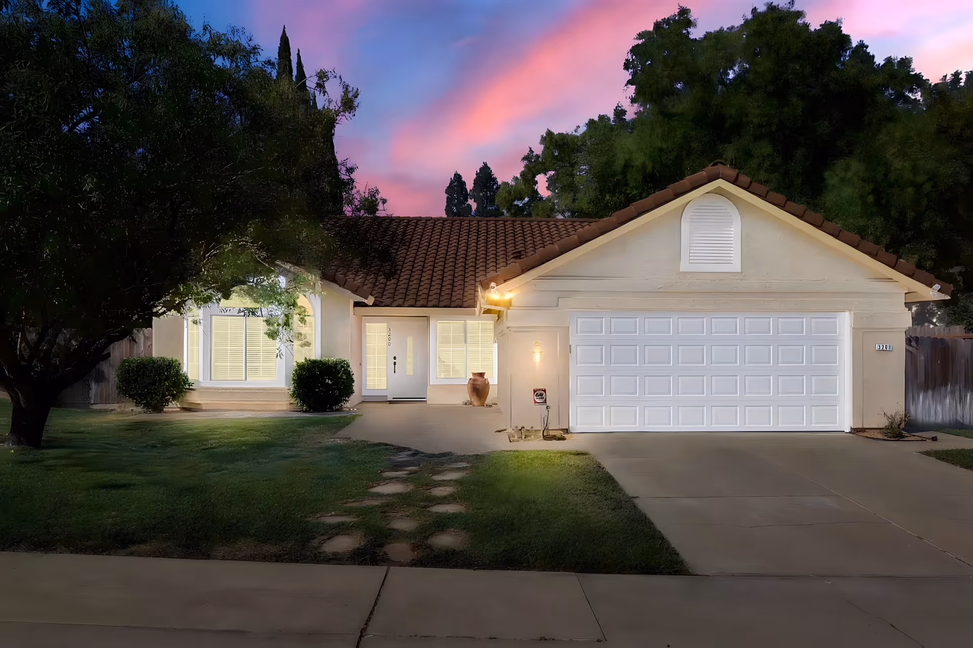 Exterior front view of a single-story house with a tiled roof, a two-car garage with a white door, a well-maintained lawn, and trees on either side. The sky is colorful with pink and purple hues during sunset or dusk.