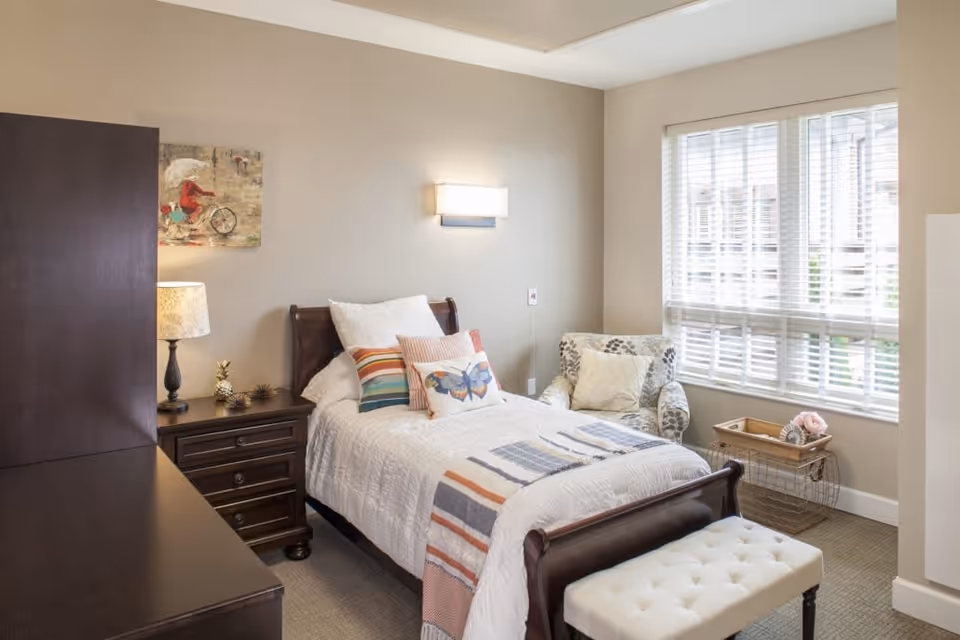 A cozy bedroom with a single wooden bed dressed in white bedding and colorful pillows, including one with a butterfly design. Next to the bed is a dark wooden nightstand with a lamp and decorative items. A patterned armchair is placed near a window with white blinds, and a small wire basket with a wooden tray and flowers sits beside it. The walls are painted beige, and a piece of artwork featuring a person on a bicycle hangs above the nightstand.