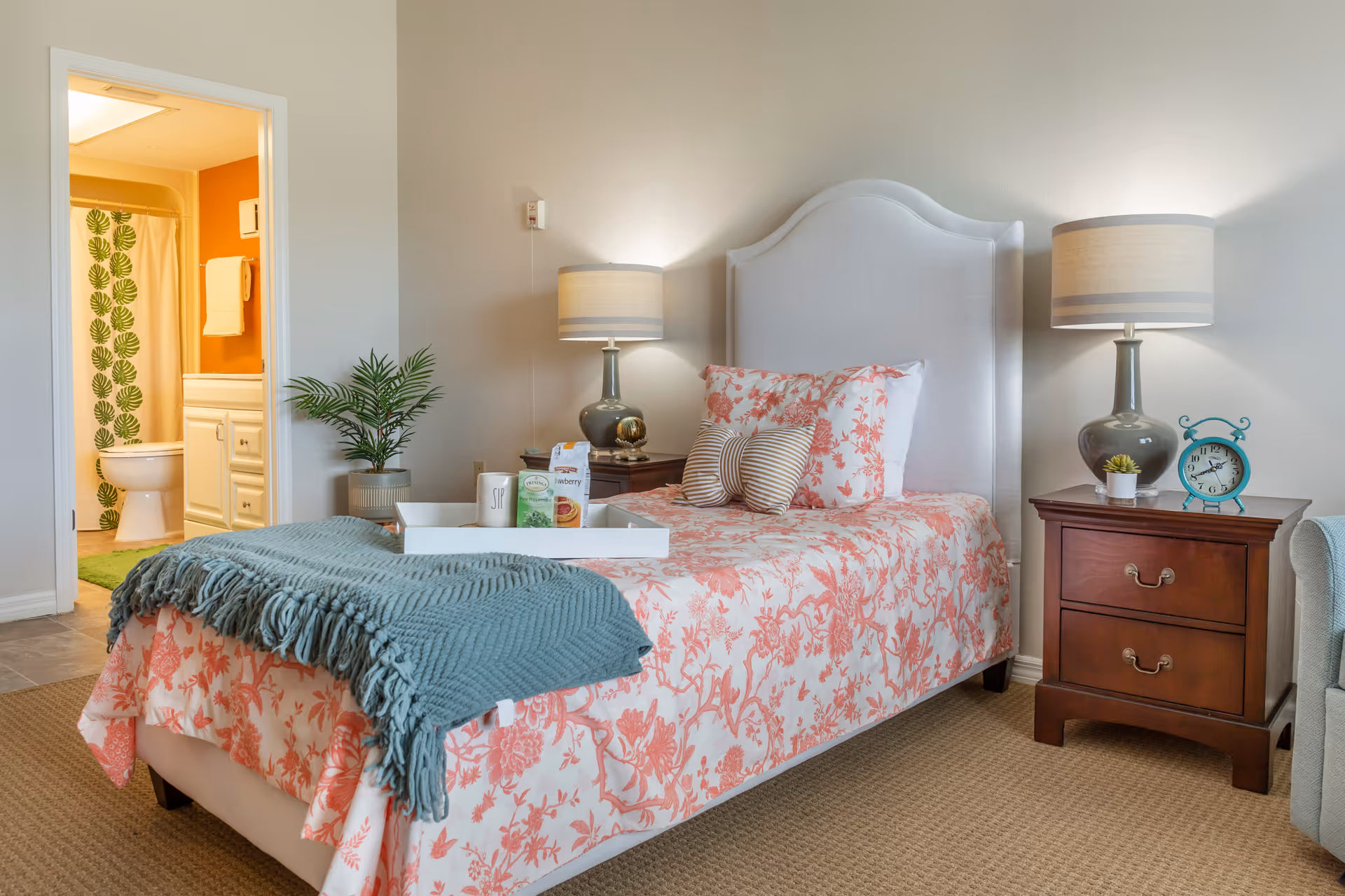 A neatly decorated bedroom with a single bed in floral bedding, bedside tables and lamps, and a view into an adjacent bathroom.