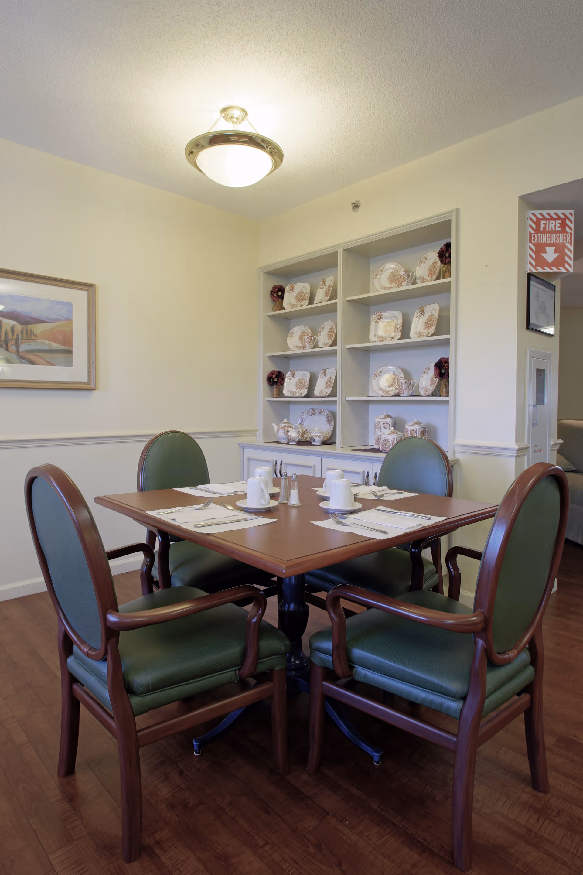 A dining area with a square wooden table set for four people with white cups, saucers, and silverware. Four green cushioned chairs with wooden frames surround the table. Behind the table is a white cabinet with shelves displaying decorative plates and teapots. A framed landscape painting hangs on the wall to the left, and a ceiling light fixture is visible above the table.