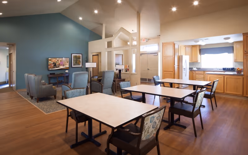 A spacious and well-lit common area in a senior living facility featuring a kitchen with wooden cabinets and a refrigerator, several tables with chairs arranged for dining or activities, and a seating area with armchairs and a television mounted on a blue accent wall.