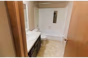 A small bathroom with a white bathtub and shower combination, a white toilet, a sink with a countertop, and a mirror above the sink. The floor has light-colored tiles, and the walls are painted white. The door to the bathroom is partially open.