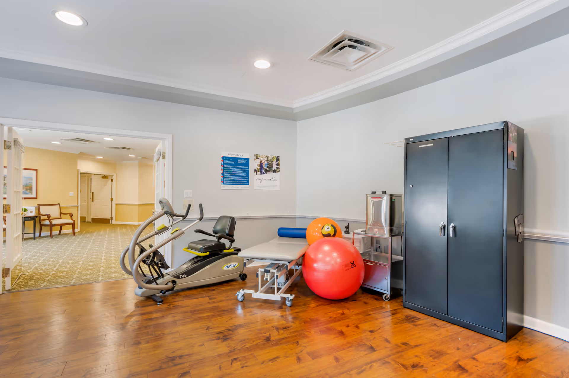 Therapy/exercise room with a recumbent bike, treatment table, large exercise balls, a black storage cabinet, and a water dispenser.