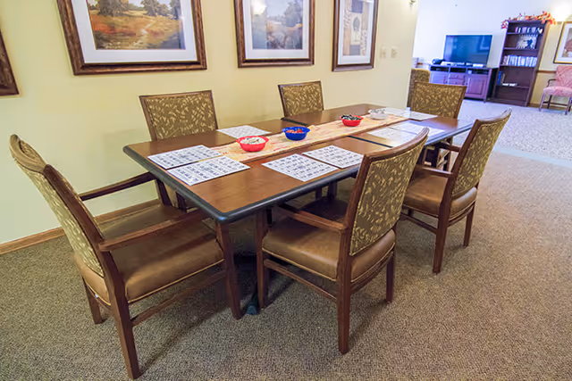 Rectangular table with six upholstered chairs set with bingo cards and bowls in a communal dining/activity room.