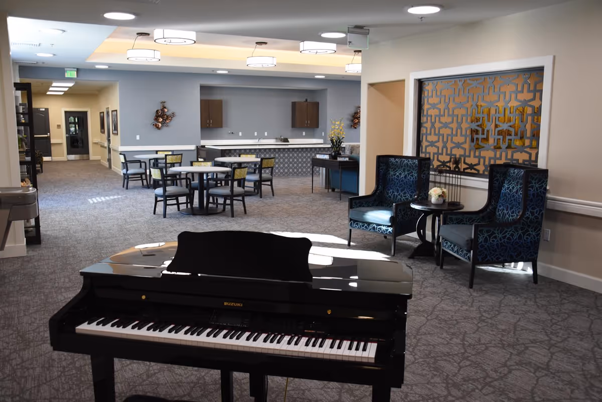 Spacious senior living common room with a black piano in the foreground, round dining tables and chairs in the center, and lounge seating by a decorative partition.