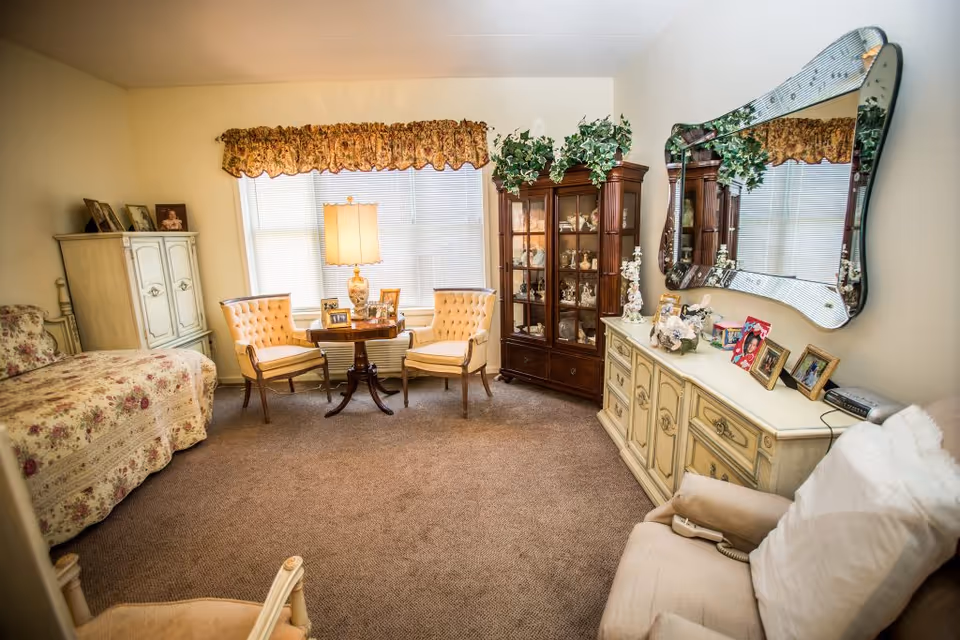 A cozy, well-lit bedroom with floral bedding on a single bed, two upholstered chairs around a small round wooden table with a lamp and framed photos, a wooden cabinet with glass doors displaying decorative items, a large decorative mirror above a cream-colored dresser adorned with more framed photos and ornaments, and a comfortable armchair with a pillow and a remote control on it.