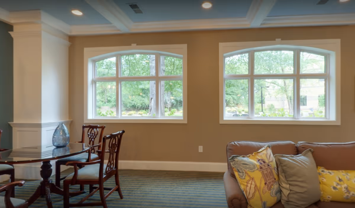 Interior view of a living room area with two large arched windows showing green trees outside. The room has a brown sofa with colorful pillows on the right and a round glass-top table with wooden chairs on the left. The walls are beige with white trim and the ceiling has recessed lighting.
