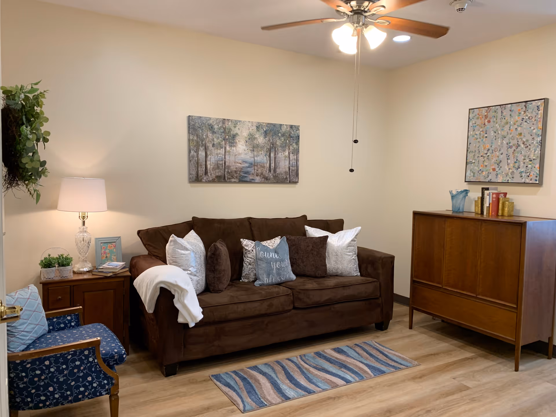 A cozy living room with a brown sofa adorned with multiple pillows, including one with the text 'Count your blessings'. There is a blue patterned armchair to the left, a wooden side table with a lamp and small plants, and a wooden cabinet on the right with decorative items and books on top. The walls are decorated with two paintings, and a ceiling fan with lights is visible above. The floor has a light wood finish with a small striped rug in front of the sofa.