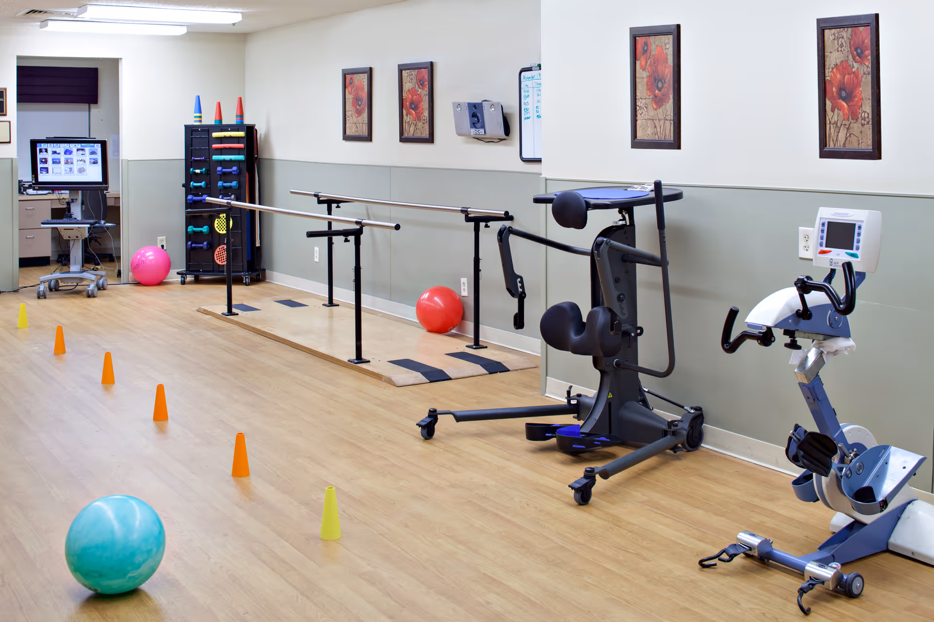 A physical therapy or rehabilitation room with parallel bars for walking exercises, exercise balls, small cones set up on the wooden floor, and various exercise equipment. The walls are decorated with framed floral artwork, and there is a computer workstation in the background.