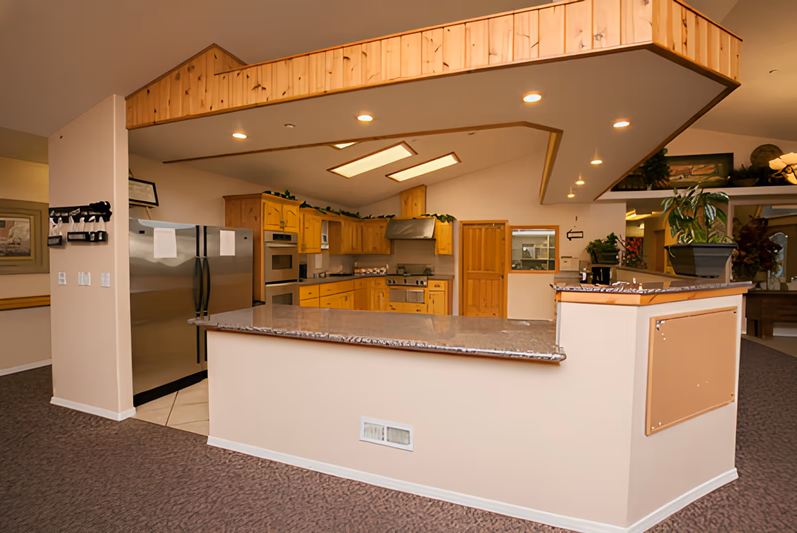 Interior view of a kitchen area in an assisted living facility featuring wooden cabinets, stainless steel refrigerator, built-in oven, stove with a range hood, and a granite countertop bar with overhead recessed lighting.