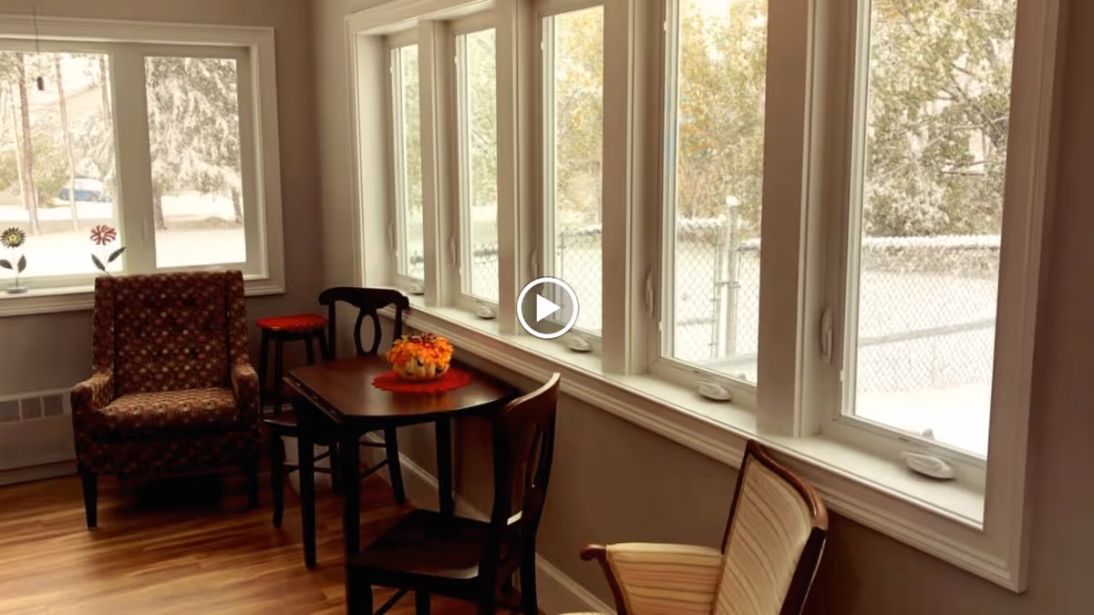A cozy sitting area with a patterned armchair, a small wooden table with a floral centerpiece, and three wooden chairs placed near a row of large windows overlooking a snowy outdoor scene.