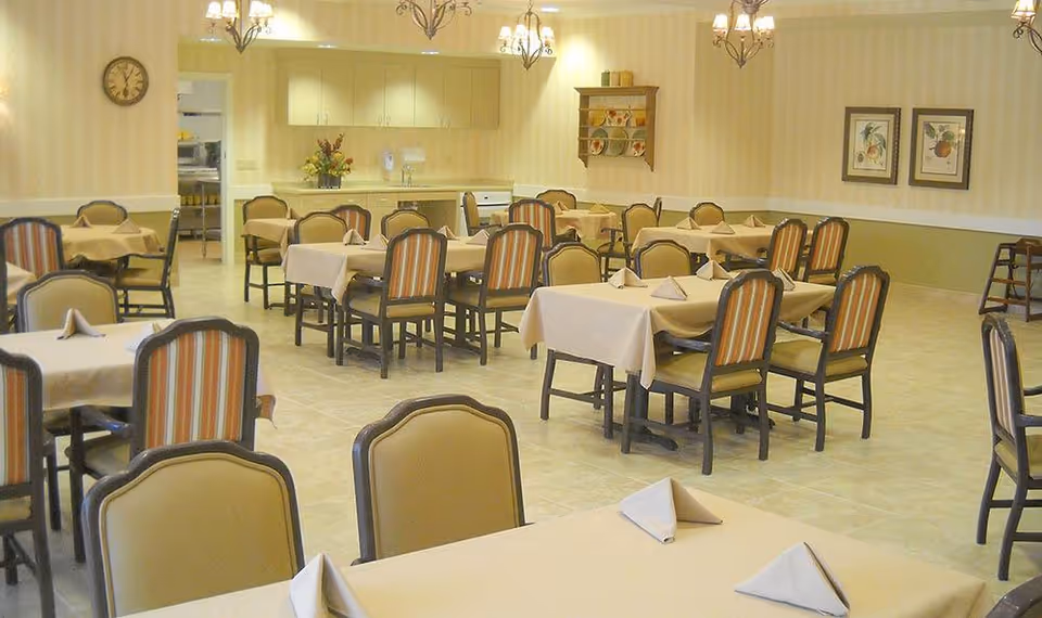 A dining room with multiple tables covered in beige tablecloths and set with folded napkins. The chairs have beige and striped upholstery. The room has tiled flooring, chandeliers, a clock on the wall, framed artwork, and a kitchen area in the background.