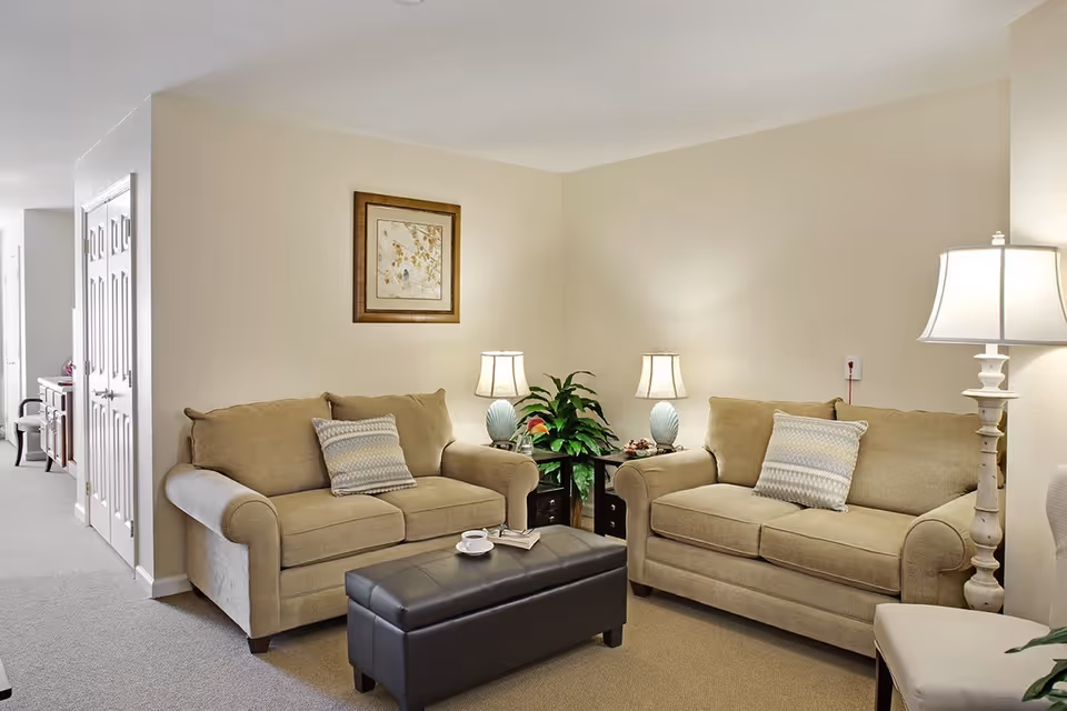 A cozy living room with two beige sofas facing each other, each with a patterned throw pillow. Between the sofas is a small dark brown ottoman with a cup and saucer on top. There are two matching table lamps on side tables behind the sofas, a framed artwork on the wall, and a green potted plant in the corner. The room has light-colored walls and carpeted flooring.