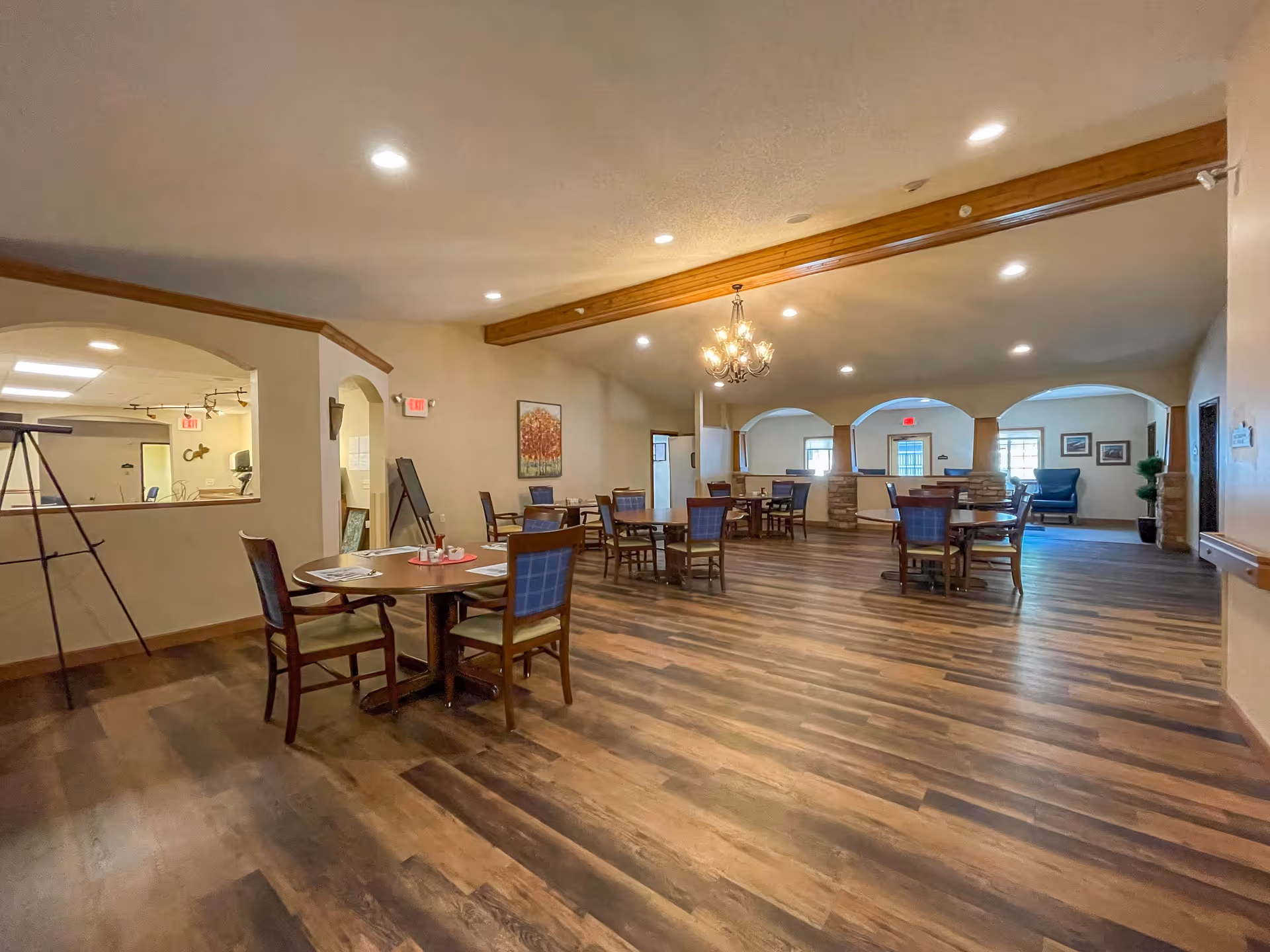 Spacious dining room with multiple round wooden tables and chairs arranged on a wood-patterned floor. The room features a chandelier hanging from the ceiling, recessed lighting, and arched openings leading to other areas. There is a painting on the wall and a comfortable armchair in the background near windows.