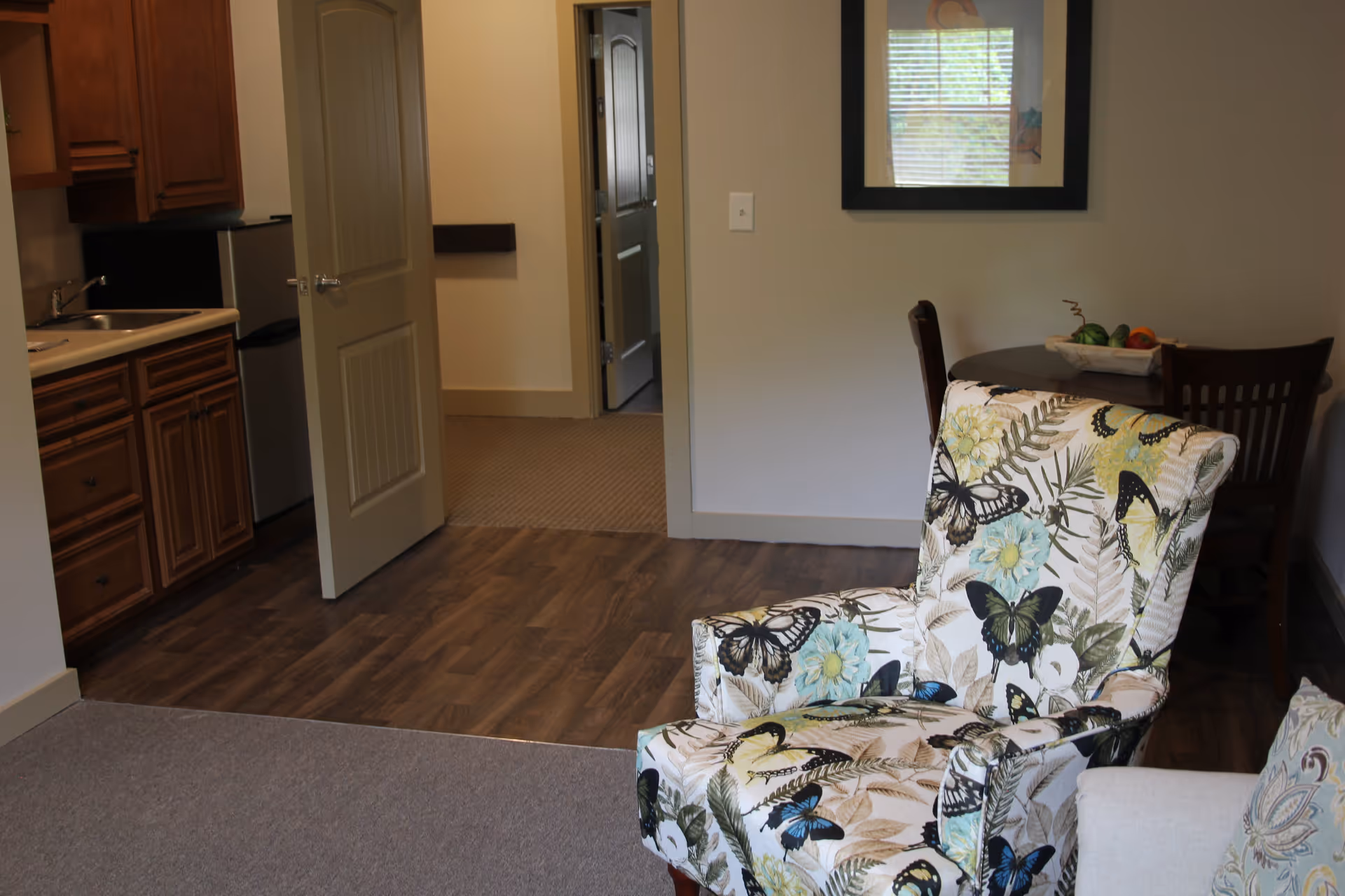 Cozy living area with a butterfly-patterned armchair, a kitchenette to the left, an open doorway to a hallway, and a small dining table.