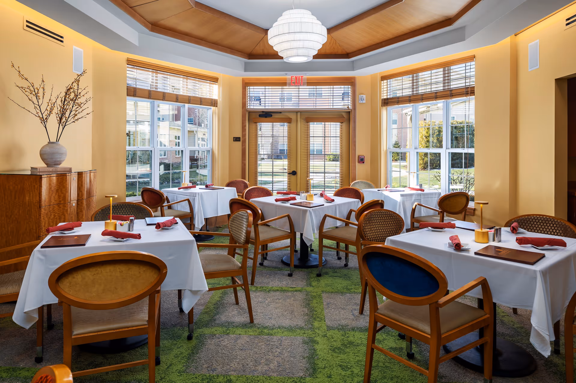A bright dining room with several tables covered in white tablecloths, each set with red napkins, menus, and small table lamps. The room has large windows with wooden blinds allowing natural light to fill the space. There are wooden chairs with cushioned seats around the tables, a wooden cabinet with a vase holding branches on the left, and a modern chandelier hanging from a wooden ceiling.